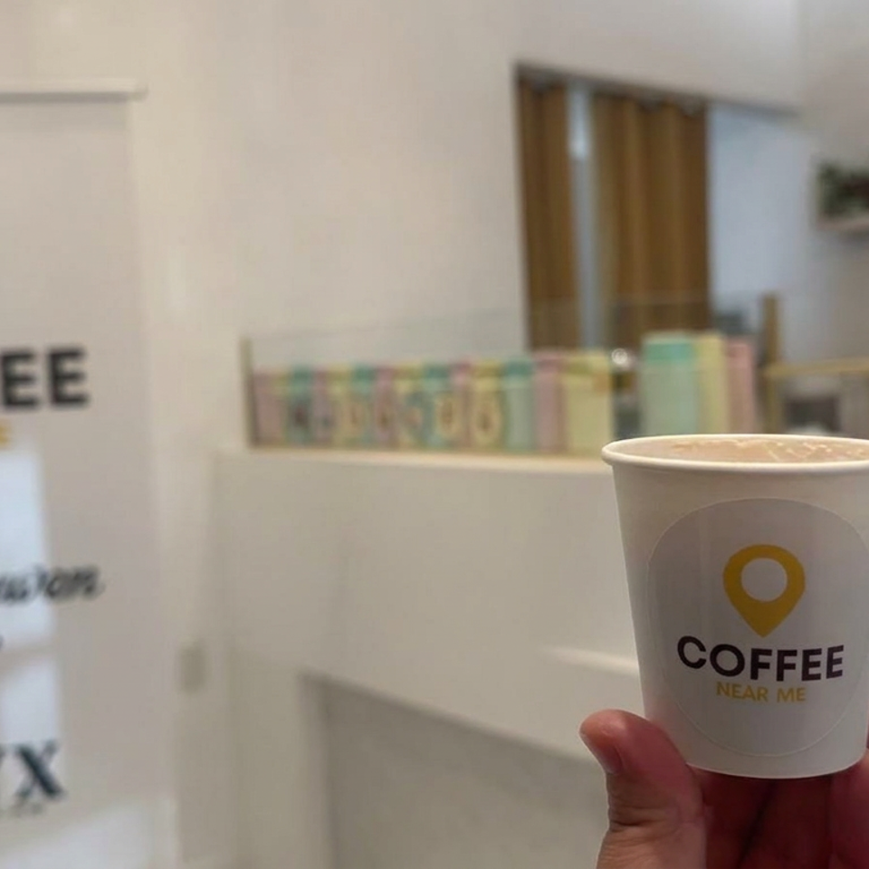 Hand holding a branded Coffee Near Me cup inside a bright café, with pastries, shelves, and a matching sign in the background.