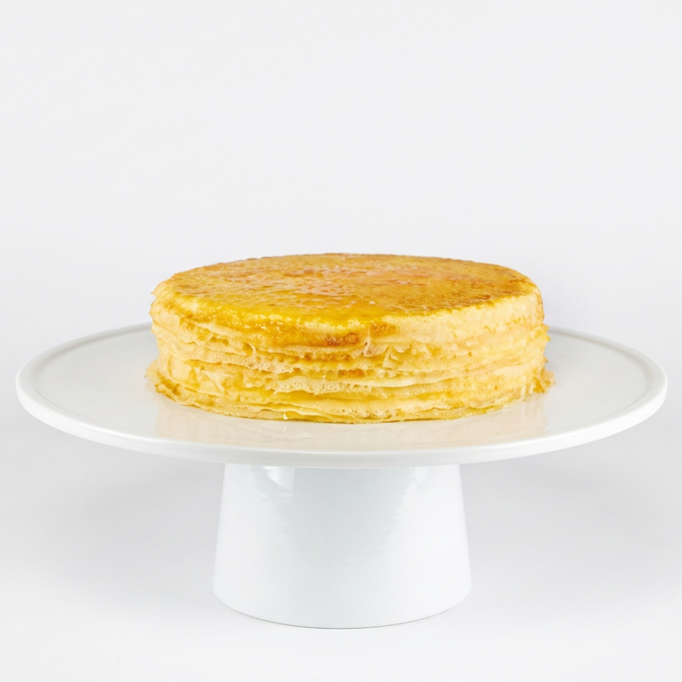 Stack of golden pancakes displayed on a white pedestal cake stand against a plain white background.